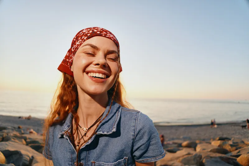 Smiling woman in modern boho-western denim street style enjoys sunset by the beach with film color effect and natural light, radiating joy and warmth.