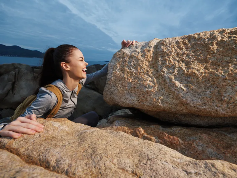 Young woman climbing rocky terrain, showcasing determination and joy against a serene coastal backdrop, wearing outdoor gear in a vibrant setting