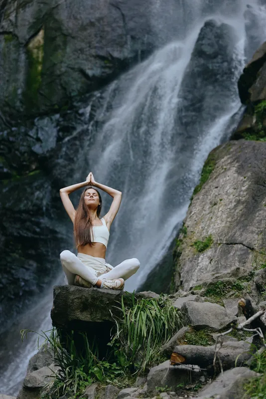 Young woman meditating outdoors near a waterfall, wearing white sportswear, practicing yoga in a serene natural environment surrounded by rocks and greenery.