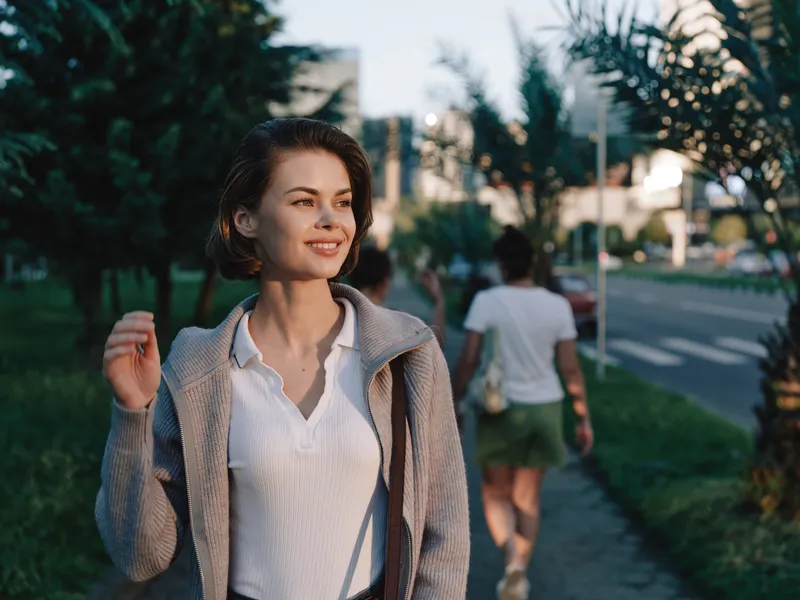 A cheerful woman walks along a sunny urban sidewalk. She smiles warmly, enjoying the moment and a calm, confident mood.