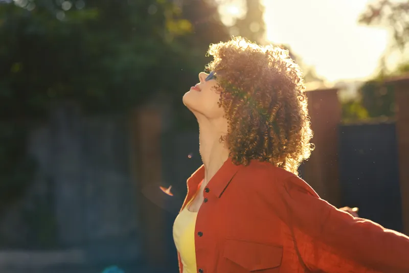 Bright young woman outdoors tilts her head back with eyes closed as sunlight pours over her, wearing a redshirt over a yellowtop, embodying a carefree sunlit moment