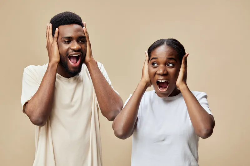 Excited young African couple expressing joy and surprise with hands on heads, wearing casual light colored clothing against a soft beige background