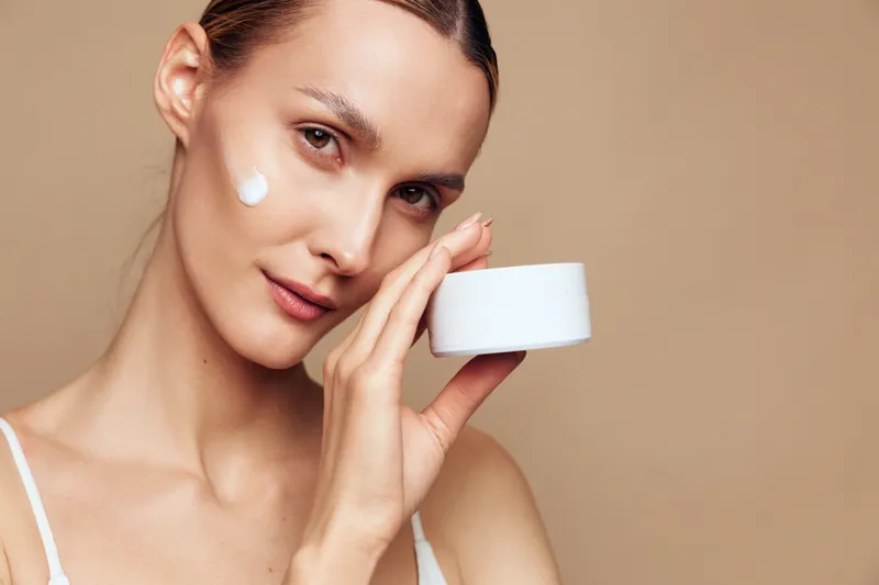 Young woman with clear skin applying cream on her face holding a white jar, skincare routine, natural beauty, soft lighting, beige background, closeup portrait