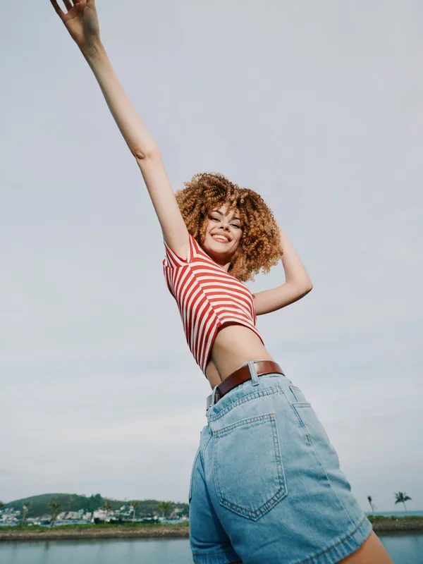 Vibrant young woman with curly hair wears a red striped top and denim jeans, raising an arm in celebration against a clear sky by the water.