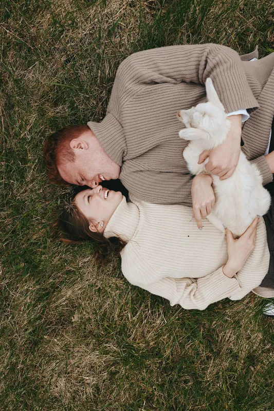 A couple lies on grass, embracing a white goat kid between them, sharing a warm, intimate moment in a calm outdoor setting. Cozy sweaters, soft light, and smiling faces convey affection, connection,