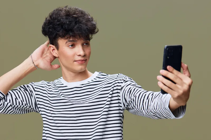 young man taking selfie with smartphone wearing striped shirt with curly hair on olive background, casual style and relaxed expression in studio lighting