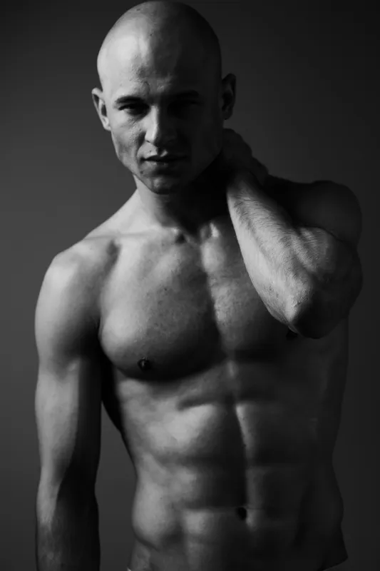 Black and white image of a muscular man posing confidently against a dark background, showcasing his physique. Ideal for fitness and health themes.