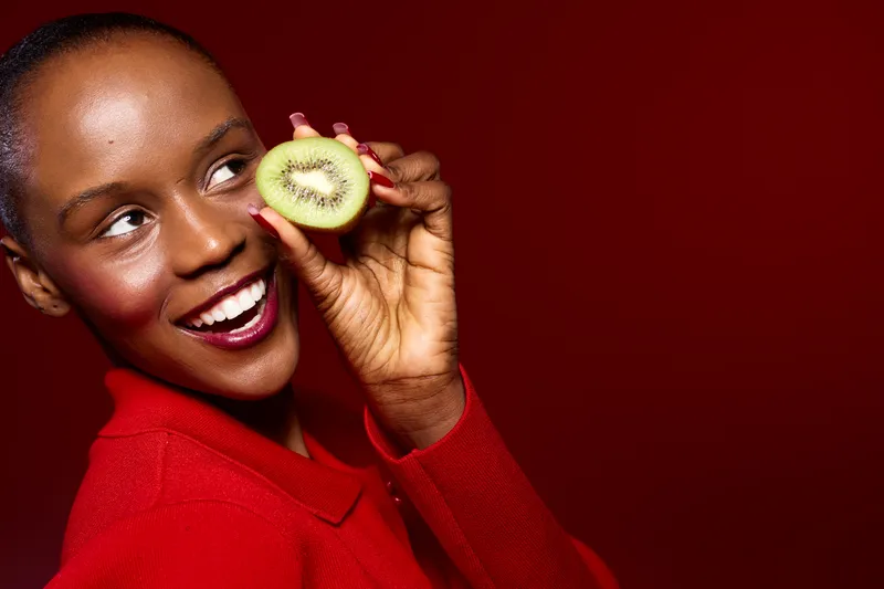 Smiling black woman with dark skin and short hair holds a slice of kiwi near her face. She wears a red sweater against a red background. Concept of healthy lifestyle and fresh fruit.