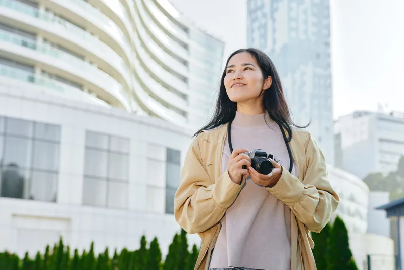 Woman with camera in urban cityscape, candid street style photography capturing exploration, daylight scene with modern buildings, greenery and travel vibe, creative storytelling through imagery
