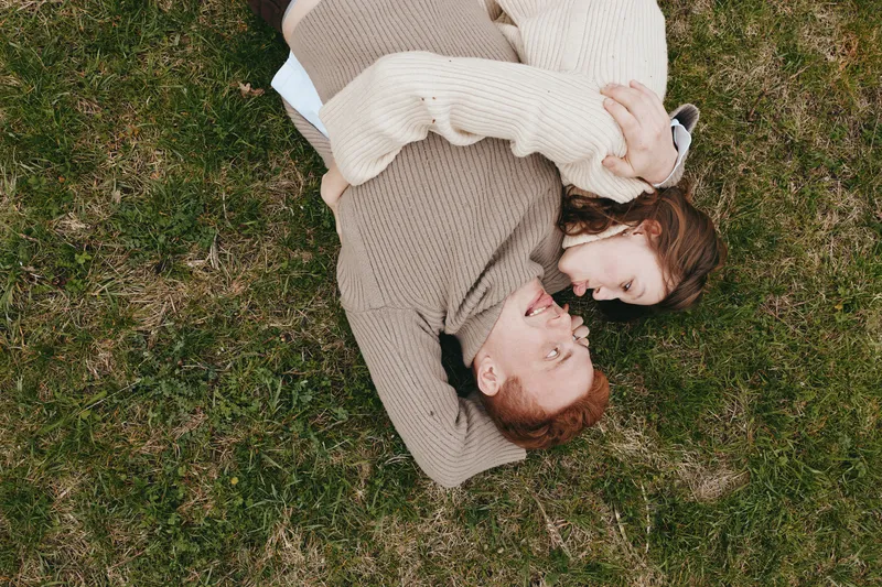 Two lovers lie on a grassy field, embracing in cozy knitwear. The intimate outdoor moment conveys romance, warmth, and a relaxed lifestyle in a bright, natural setting.