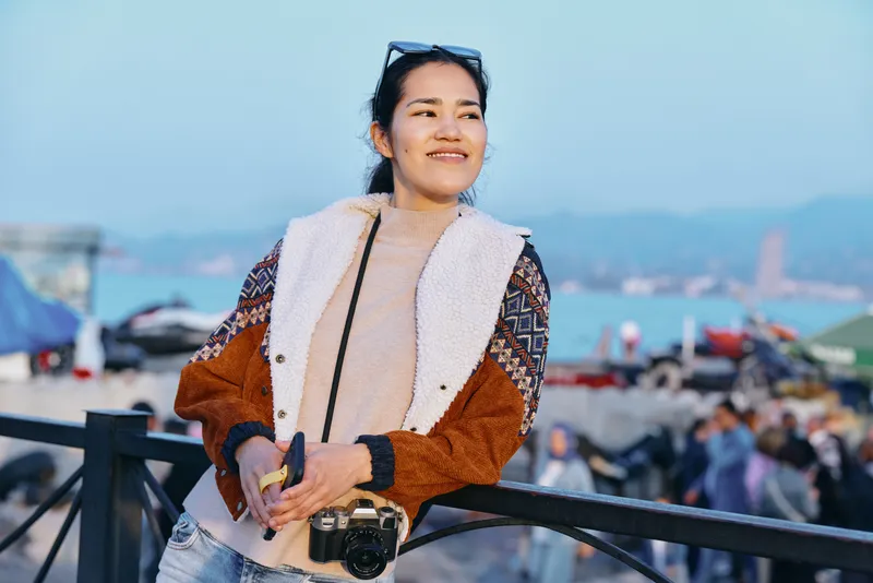 Woman leaning on a railing with a camera, wearing a plush brown faux fur jacket, by the water with a coastal backdrop during a sunny travel day