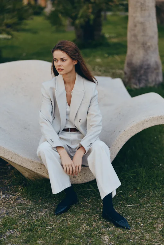 confident woman in white suit sitting outdoors on unique stone bench with green grass and trees in fashion business concept
