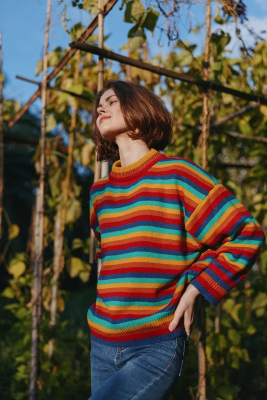 Woman in mid 20s with short hair wearing a colorful striped sweater and casual outfit, smiling and leaning in outdoor garden under warm autumn light, lifestyle portrait.