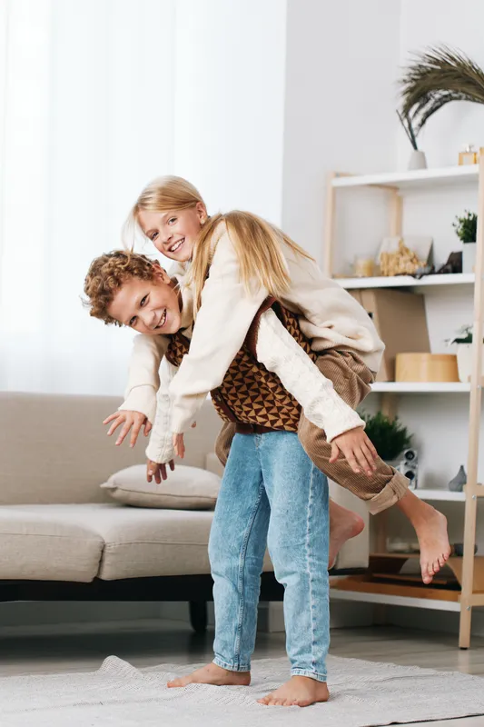 Playful couple enjoys a light moment at home, one person carries the other on their back, smiling, casual attire, cozy living room, warm light, relaxed joyful mood.
