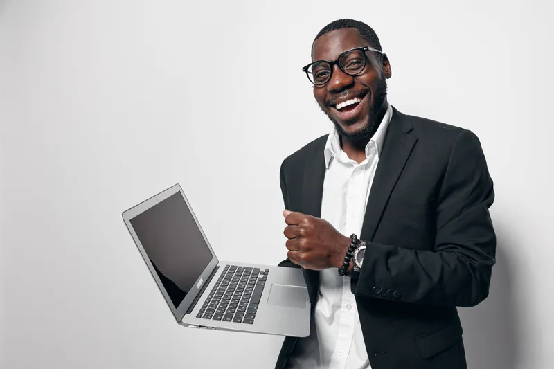 African American man with dark skin wearing glasses and black suit holding laptop and smiling confidently in studio. Business professional, technology, success and positive emotion.