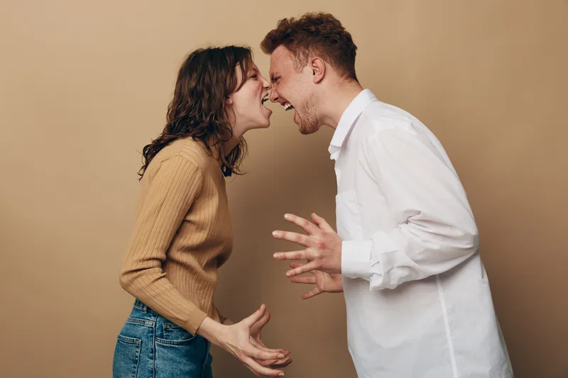 Angry young man and woman shouting at each other in confrontation. Casual clothing, emotional conflict, and relationship tension on beige background. Concept of argument and disagreement.