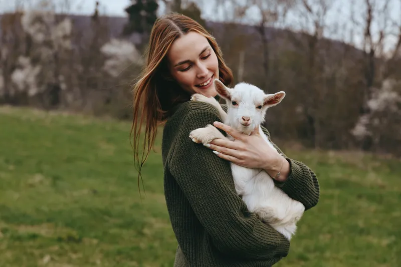 Cheerful woman outdoors, embracing a small goat kid, cozy sweater, gentle breeze, sunlit moment, warm smile, lush greenery background, casual lifestyle scene showing harmony with animals.