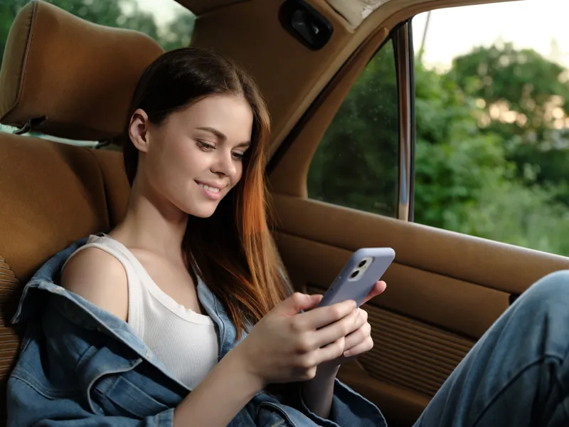 Young female model enjoying her smartphone in a car, wearing casual denim attire and displaying a joyful expression, surrounded by lush greenery.