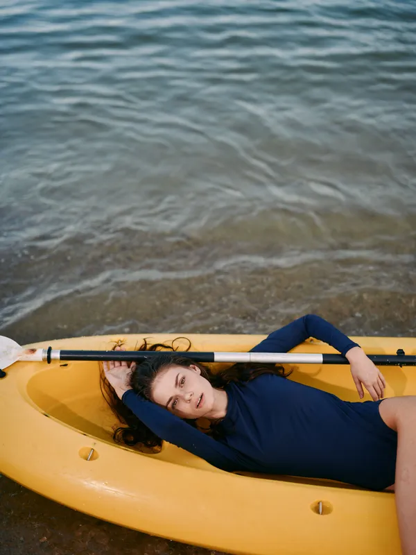 Relaxed woman lying on yellow kayak by water shore in blue swimsuit with calm expression and natural background.