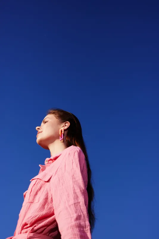 A young woman of European descent wearing a pink shirt against a clear blue sky, showcasing confidence and tranquility in a summer setting