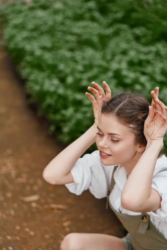 Young woman in a garden, playfully posing with hands on her head, wearing casual summer attire, surrounded by lush greenery, smiling and enjoying the moment