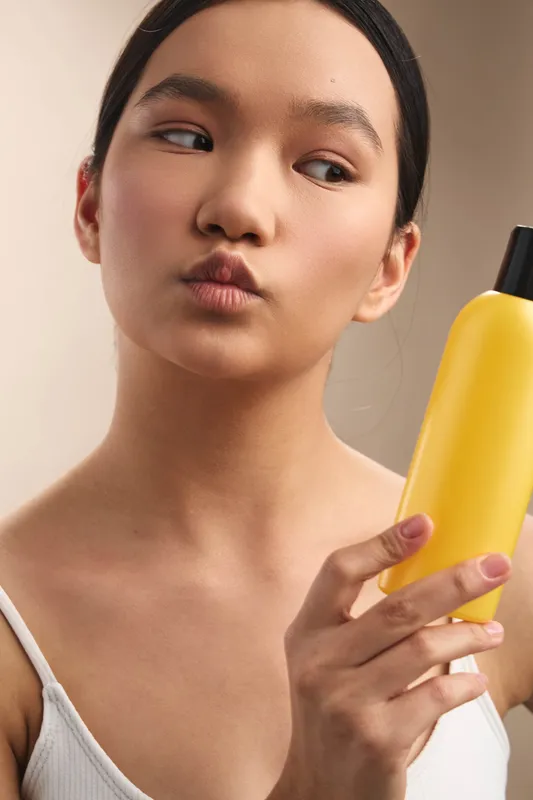 Asian woman holds a yellow bottle near her face, with a thoughtful expression and soft lighting, presenting a calm beauty moment in a clean indoor setting.