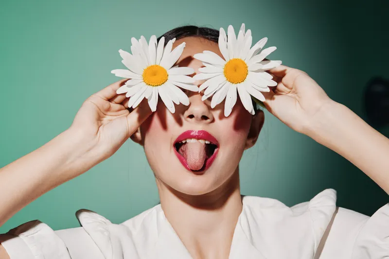 Playful young woman with glossy lips holds two white daisies over her eyes sticking out her tongue dramatically. Bright green backdrop and white shirt highlight queen concept dreams.