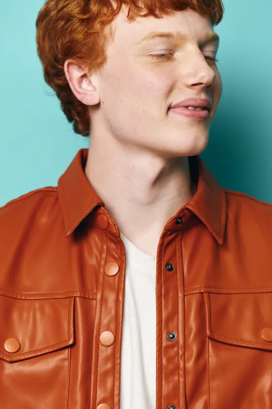 Young man with red hair and freckled skin wearing brown leather jacket and white t shirt eyes closed on blue background. Portrait of peaceful male with stylish casual look and calm expression.