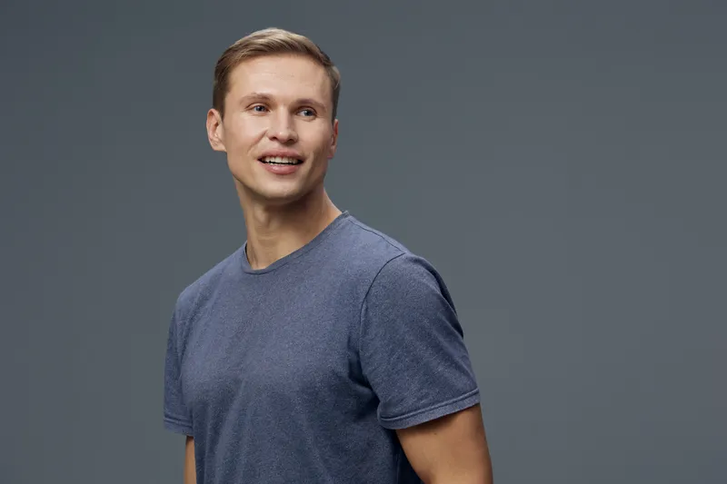 Smiling man in casual blue t-shirt against plain gray background, looking to the side, happy and relaxed, studio portrait, lifestyle concept, positive emotions, natural appearance, and modern style.