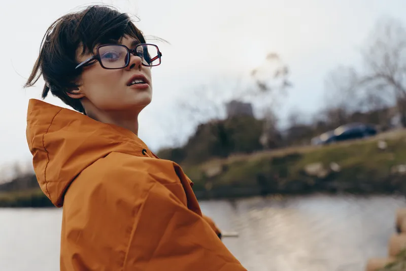 woman lifestyle outdoors near river lake, orange jacket and glasses, close portrait profile, windy day with hair blowing, she looks away with thoughtful expression, soft nature background