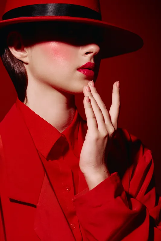 Fashionable woman in red coat and wide hat softly touches face, showing bold red lips and flawless skin under dramatic shadow. Concept of beauty, fashion, and elegance.