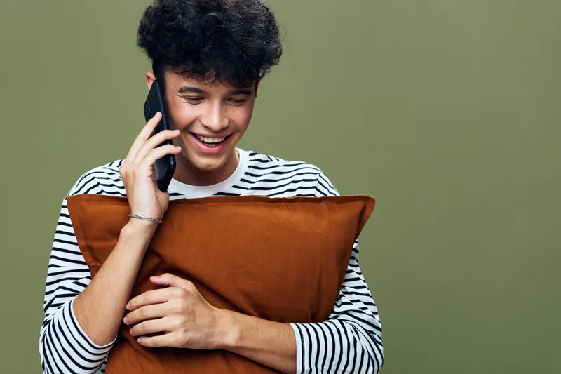 smiling young man talking on phone holding brown pillow against olive background, casual striped shirt, happy expression, indoor portrait.