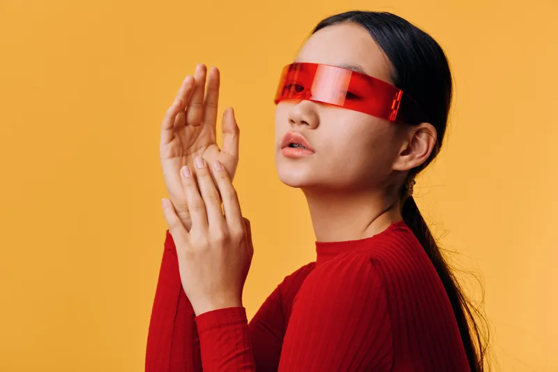 Woman with long dark hair wearing red futuristic glasses and matching red sweater posing with hands near face against orange background. Fashion, style, modern look, technology, female model.