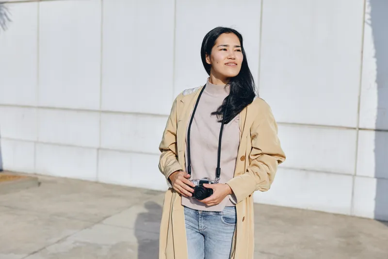 Woman outdoors in a beige trench coat holding a camera, casual confident pose, light sweater and jeans, sunny day by a modern wall, lifestyle photography and travel vibe