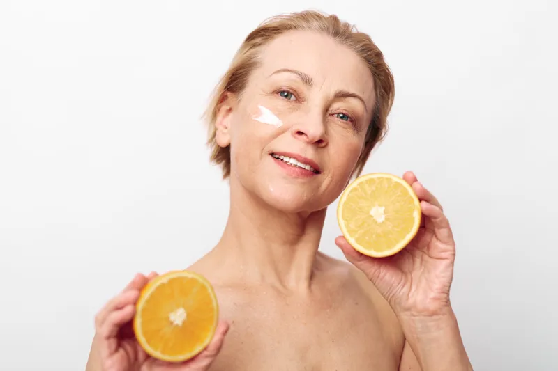 Mature woman with natural skin holding two halves of an orange applying facial cream, isolated on white background. Concept of skincare, natural beauty, health, and wellness.