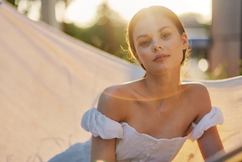 Young woman in white dress relaxing in hammock, enjoying sunlit nature, showcasing serene emotions and summer vibes