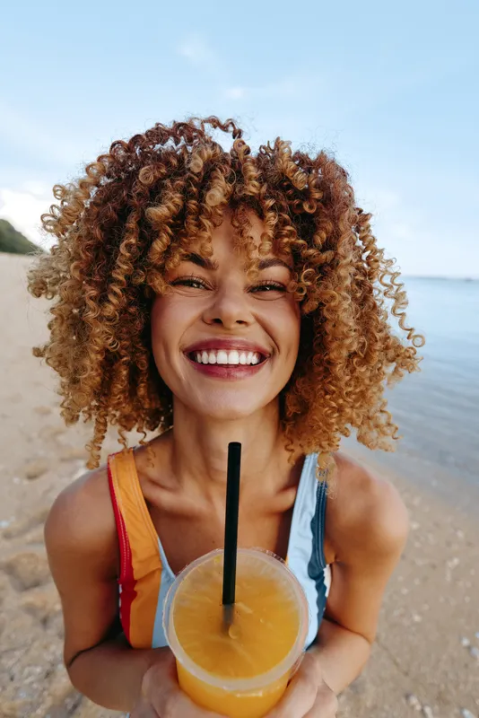 A happy young woman with curly hair smiles brightly while holding a fresh orange juice on a sunny beach. The clear sky and calm sea create a joyful summer vibe.