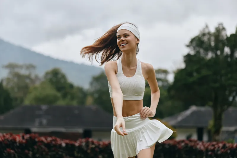 Young woman in white sportswear joyfully running outdoors, surrounded by nature, showcasing an active lifestyle and summer fitness enthusiasm