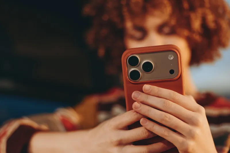 Woman lifestyle selfie indoors, smartphone with brown case in hand, phone close view, curly hair blurred, cozy home bedroom portrait, warm soft light
