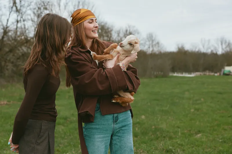 Smiling woman wearing a beanie holds a playful goat kid in a sunny field, while another woman watches. A candid lifestyle moment showing bonding with animals, outdoor adventure, and countryside charm.