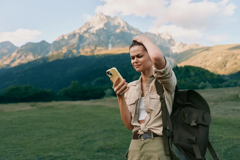 A young woman with a backpack stands outdoors in front of mountains, looking confused while holding a smartphone. She wears casual clothes in a natural landscape setting.