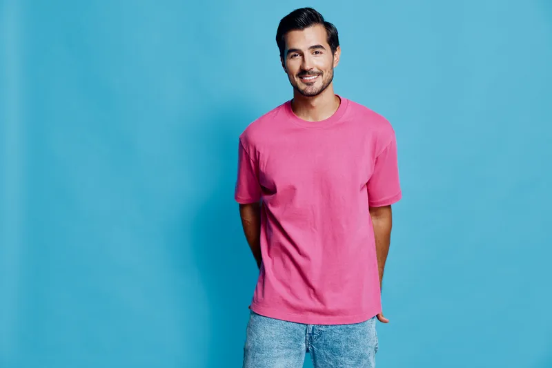 Full body smiling happy cheerful young caucasian man wear pink t-shirt stand hands in pockets isolated on solid bright blue background studio portrait. People lifestyle concept