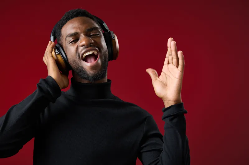 Young Black man enjoying music with headphones, wearing a black sweater against a red backdrop, expressing happiness and excitement through music