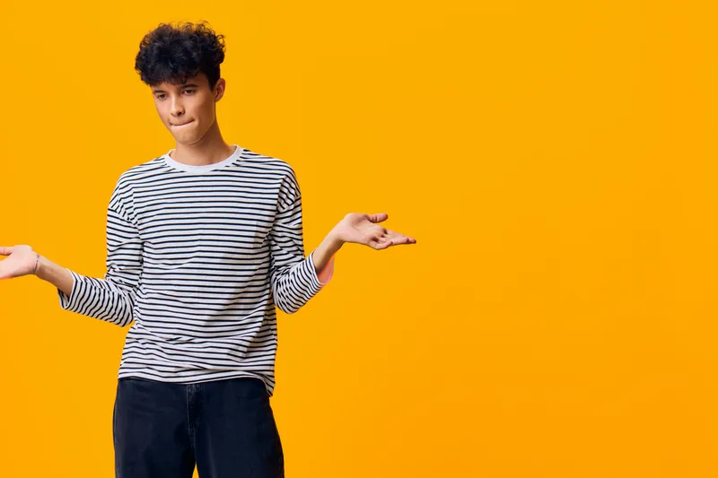 Young man with curly hair in striped shirt stands confused with open palms on vivid orange background, showing doubt and uncertainty in casual style.