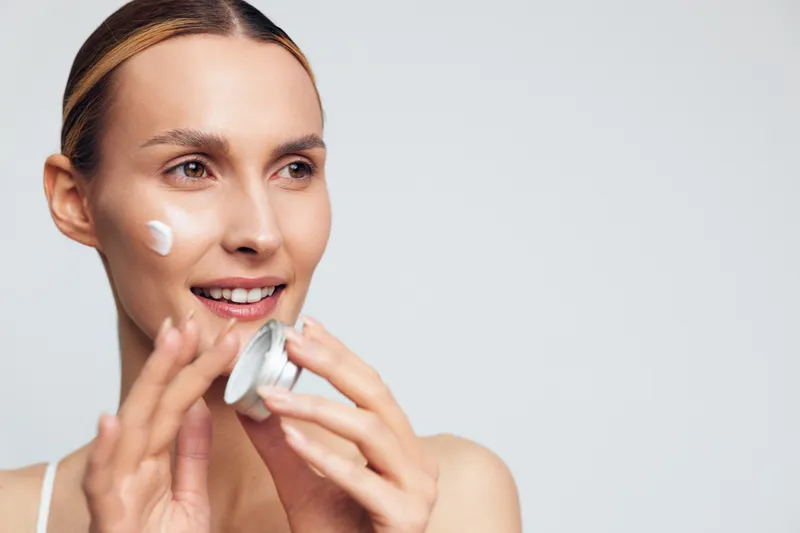 Young woman applies face cream on cheek smiling with natural skin care routine isolated on white background holding jar of moisturizer in hand with fresh healthy look.