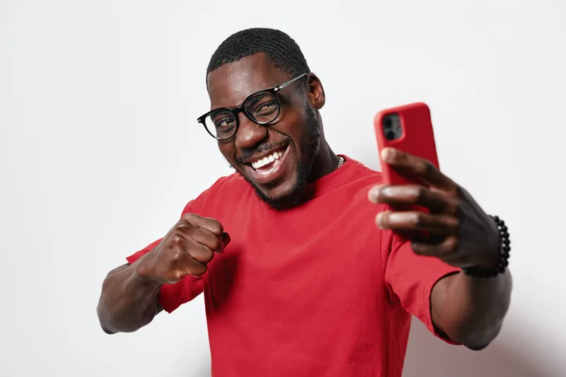 African American man with dark skin wearing glasses and red shirt taking selfie with smartphone, smiling and showing fist gesture in studio with white background and positive expression.