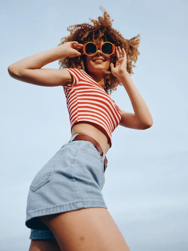 A carefree young woman wearing a red and white striped crop top and denim shorts smiles while posing against a clear blue sky, wearing bold sunglasses.