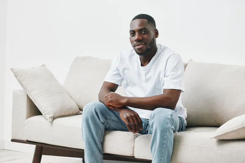 Happy young Black man in casual white t shirt and jeans sitting on a modern sofa, smiling confidently in a bright, minimalistic living room setting