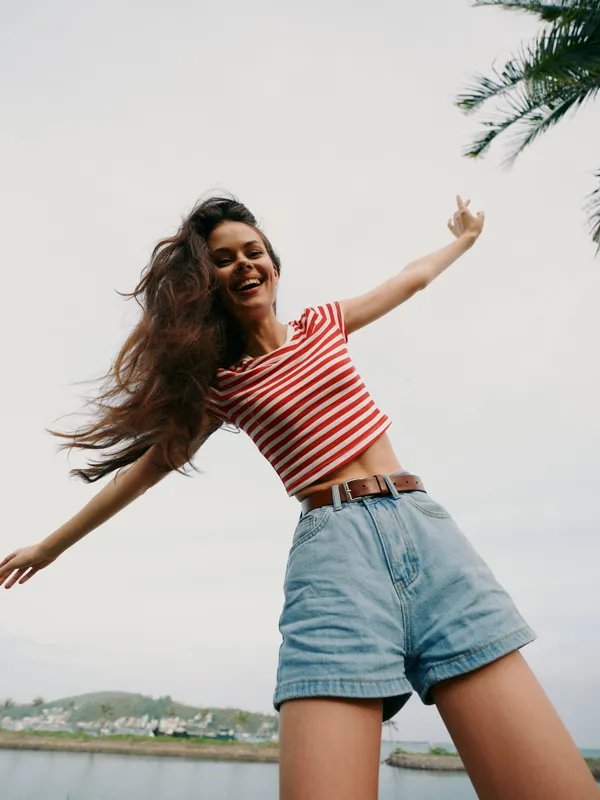 Joyful young woman outdoors with arms outstretched, wearing a red striped top and denim shorts, conveying carefree energy, sunny sky, breeze, and playful motion.