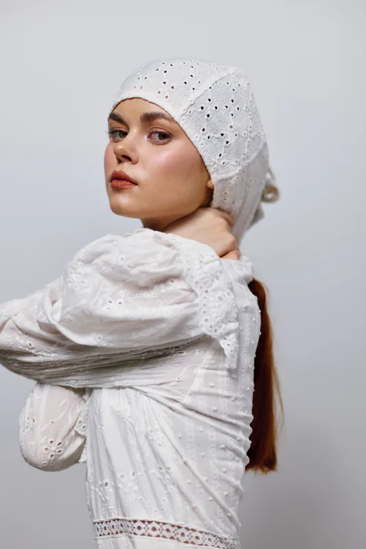 Young woman in a white embroidered dress and headscarf, expressing confidence and elegance in a minimalist studio setting with a soft color palette